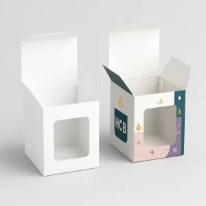 Two candle packaging boxes with open lids, one plain white and one custom printed with teal, cream, and pink design featuring 'Hi HCB' logo, candle flame, and floral illustrations, both with transparent window cutouts.