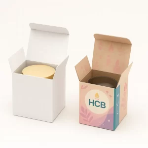 Two cube-shaped candle boxes with tuck-top lids, one plain white and one custom printed in pastel pink with teal accent, gold HCB logo, and candle flame leaf design.