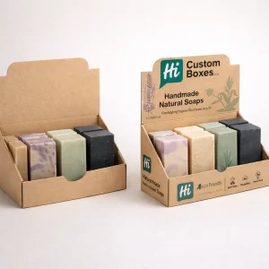 Two cardboard counter-top display units filled with assorted handmade soap bars, featuring green branding and natural botanical illustrations.