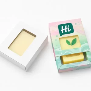 Two soap sleeve boxes- one plain white and one custom printed in pastel green and pink with leaf graphics- both with rectangular transparent windows showing the soap inside