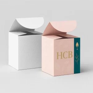 Two cube-shaped packaging boxes with round flap tops- one plain white and the other pastel pink with teal side panel- The designed box features the gold letters -HCB- with a small flame and leaf graphic